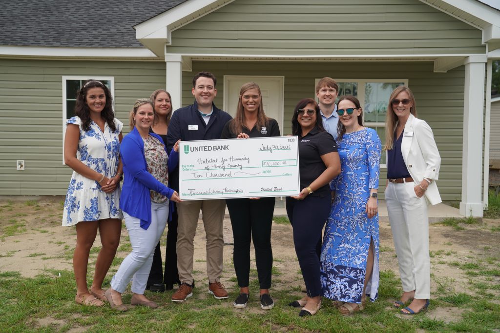 A group of nine people, dressed in business casual attire, stand in front of a newly built light green house. In the center, two individuals from United Bank hold a large novelty check for $10,000 made out to "Habitat for Humanity of Horry County" for their "Financial Literacy Partnership." The check is dated July 30, 2025. The group appears happy and engaged, representing both organizations in a collaborative event. The foreground shows some grass and dirt, indicating a recently completed construction.