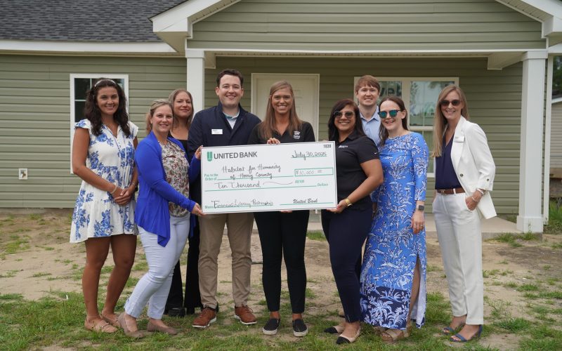 A group of nine people, dressed in business casual attire, stand in front of a newly built light green house. In the center, two individuals from United Bank hold a large novelty check for $10,000 made out to "Habitat for Humanity of Horry County" for their "Financial Literacy Partnership." The check is dated July 30, 2025. The group appears happy and engaged, representing both organizations in a collaborative event. The foreground shows some grass and dirt, indicating a recently completed construction.